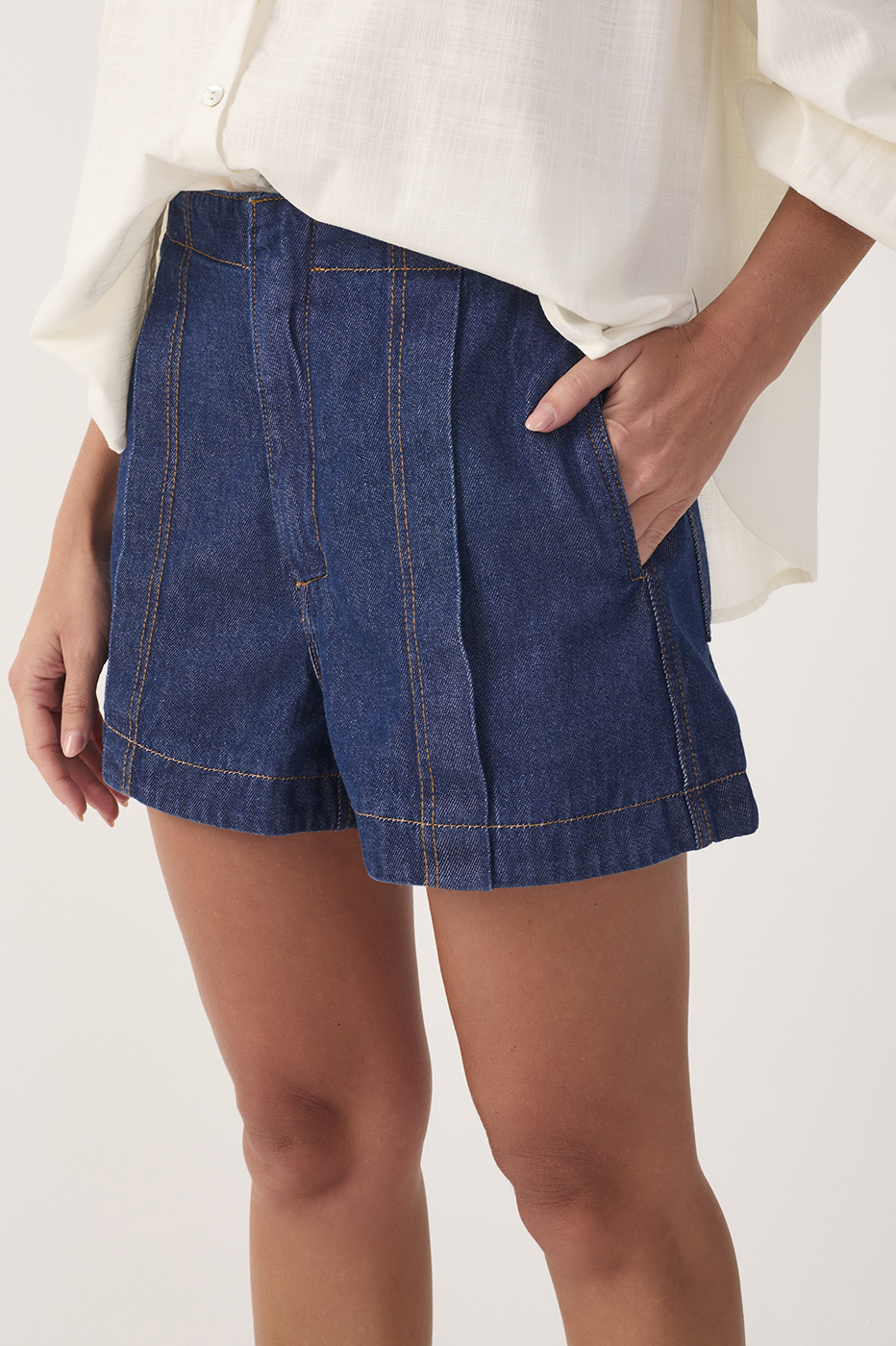 Close-up front view of dark blue denim topstitching shorts showing front pockets and orange contrast stitching, model with hand in pocket