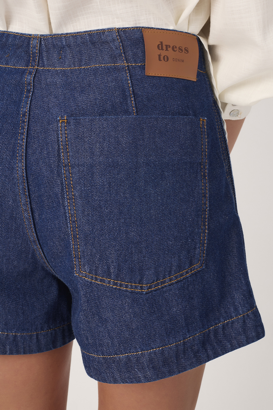 Close-up back view of dark blue denim topstitching shorts showing Dress To brand label, back pocket, and orange contrast stitching details