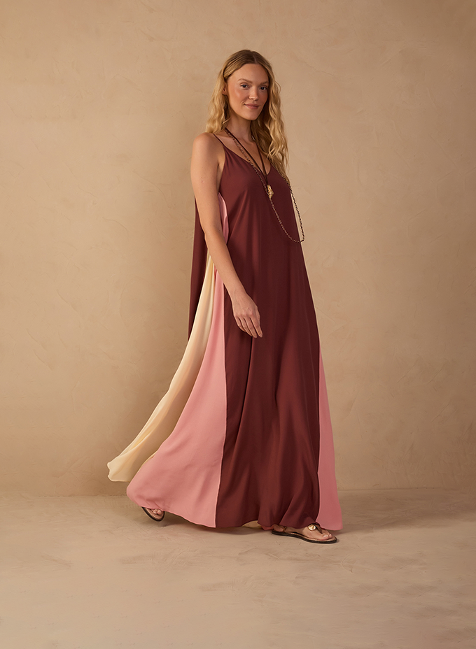 A woman stands against a warm beige textured backdrop wearing a flowing sleeveless maxi dress in deep brown with soft pink and cream side panels. The dress moves gently as she steps forward, paired with flat sandals and a long delicate necklace, creating an elegant, relaxed silhouette.