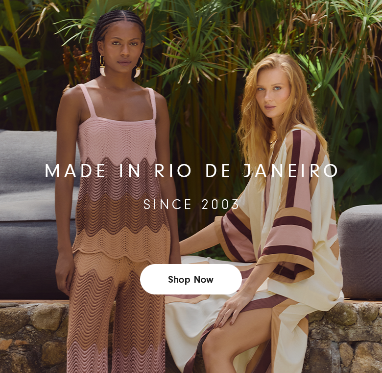 Two women pose outdoors in front of lush tropical plants and neutral lounge seating. One stands wearing a sleeveless knit outfit in blush pink, beige, and brown with a wave pattern. The other sits on a stone edge wearing a flowing cream dress with wide sleeves and pink and brown stripes. Centered over the image is the text “MADE IN RIO DE JANEIRO” and below it “SINCE 2003.”
