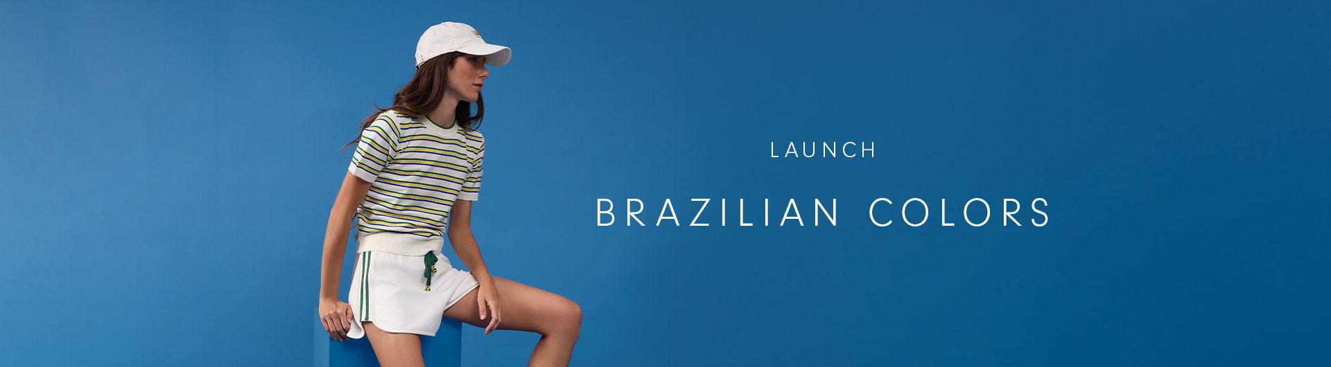 Wide desktop launch banner for the Brazilian Colors collection, featuring a woman in a white cap, striped knit top, and white shorts with green stripes leaning against a blue cube against a blue background.