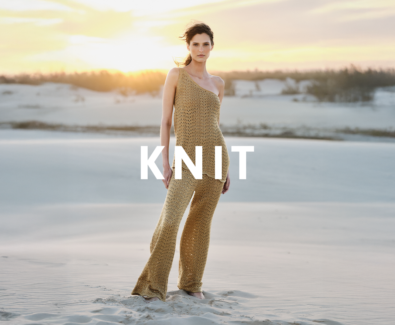 Catalog banner featuring a model in a gold, one-shoulder knit jumpsuit, with the word 'KNIT' overlaid. Setting is a sandy dune at sunset.
