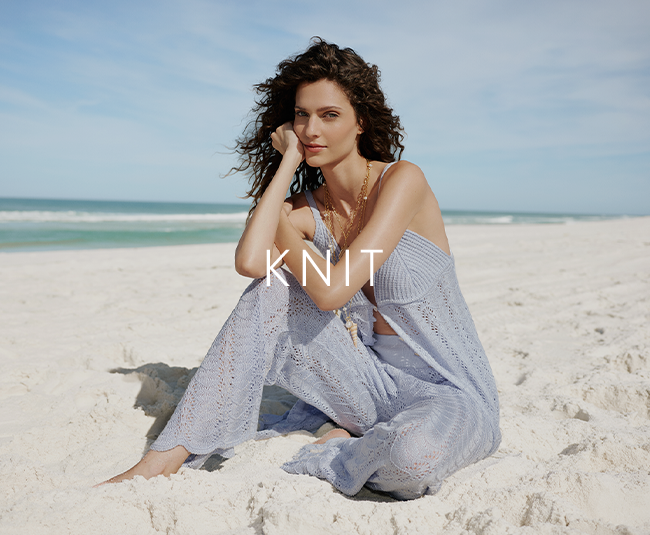 A woman sits barefoot on light-colored sand at a beach, facing the camera. She wears a light knit two-piece outfit with wide-leg pants and a matching top, featuring an open-knit texture and soft drape. The background shows a calm ocean with gentle waves and a pale blue sky. Centered over the image, the word “KNIT” appears in white, uppercase letters.