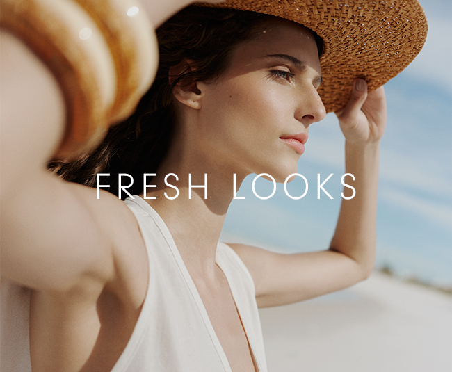 Close-up side view of a woman outdoors under natural light, looking toward the horizon. She wears a light-colored sleeveless top and a woven straw hat, holding the brim with one hand. Her other arm is raised in the foreground, partially out of focus, with stacked rounded bracelets visible on her wrist. Her hair is dark and tucked behind her ear, and her expression is calm and contemplative. The background shows a soft blue sky with thin clouds and a pale, sandy landscape. Centered across the image, the text